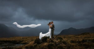 A bride and groom embrace on a grassy hilltop under a dramatic, cloudy sky. The bride’s veil is blowing in the wind, creating a flowing shape behind them. Rolling hills and mountains form the moody background.