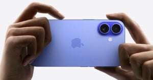 A pair of hands holding a purple iPhone horizontally. The phone has dual rear cameras and an Apple logo on the back. The background is white, emphasizing the phone's color and design.