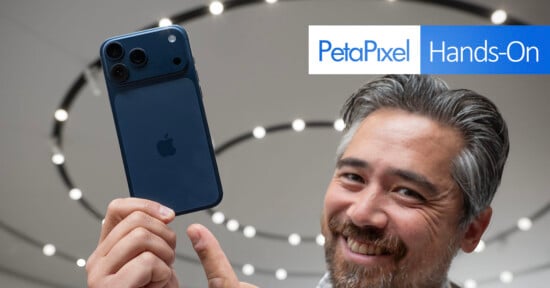 A smiling man holds up a blue iPhone with three cameras; the background features circular lights and a "PetaPixel Hands-On" sign.
