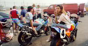 A woman sits on a colorful motorcycle, looking at the camera, while several people gather around her and another rider on a motorcycle marked "190" in a parking lot near a red van. Racing and casual atmosphere.