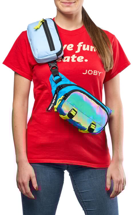 A woman wearing a red "we fun late. JOBY" t-shirt and blue jeans carries two crossbody bags, one light blue and one shiny iridescent blue, over her shoulder and across her torso.