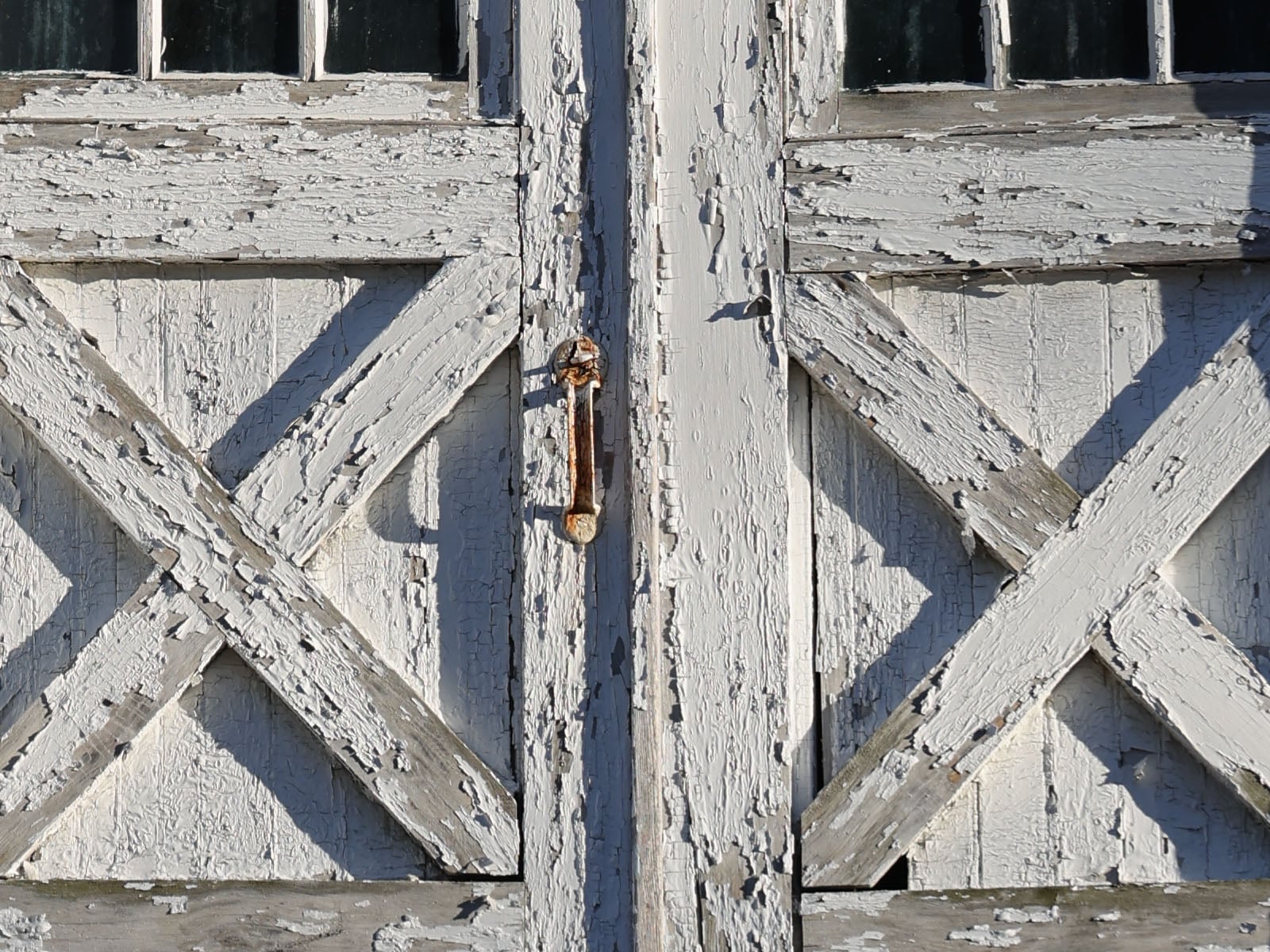 Close-up of a weathered wooden door with peeling white paint, featuring a rusty metal handle in the center and diagonal crossbeam patterns on both sides.
