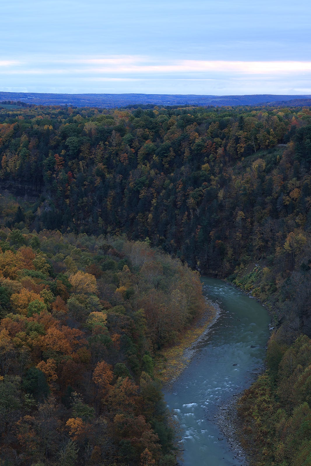 A winding river flows through a deep, tree-covered gorge with autumn foliage in shades of orange, yellow, and green under a cloudy sky.