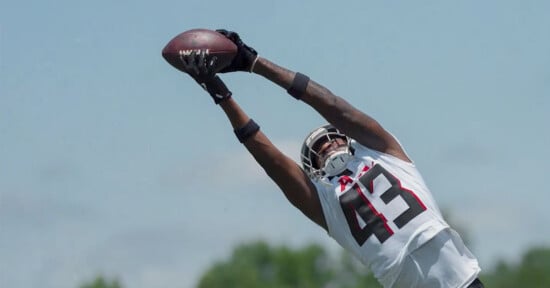 A football player in a white jersey with the number 43 stretches his arms overhead to catch a football during a practice, with a blue sky and trees in the background.