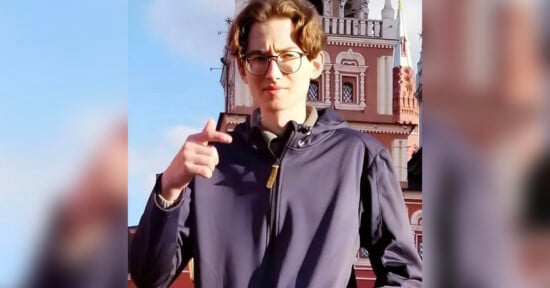 A person with wavy hair and eyeglasses is standing outside in front of an ornate building. They are wearing a dark jacket and holding up their right hand, pointing with their index finger. The building features detailed architectural designs and a partly cloudy sky.