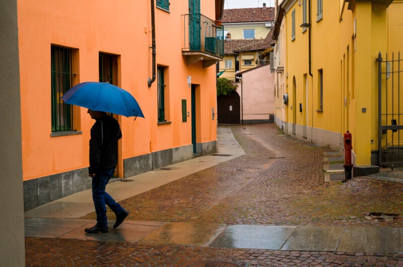 A person holding a blue umbrella walks down a wet, narrow cobblestone street lined with colorful orange and yellow buildings on a rainy day.