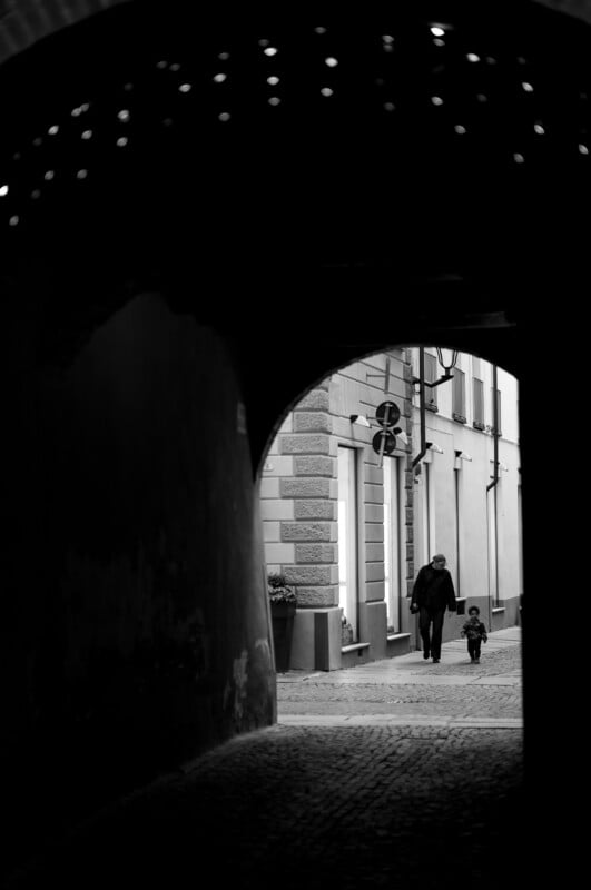 Black and white photo of an adult and a child holding hands, walking down a cobblestone street seen through a dark archway, with light illuminating the buildings ahead.