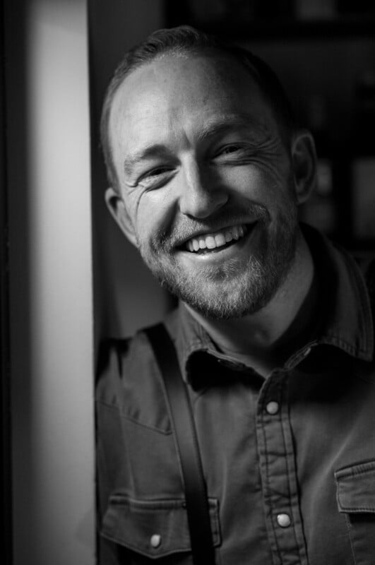 A man with a beard and short hair smiles broadly while leaning against a wall. He wears a button-up shirt and suspenders. The photo is in black and white, with soft lighting highlighting his face.