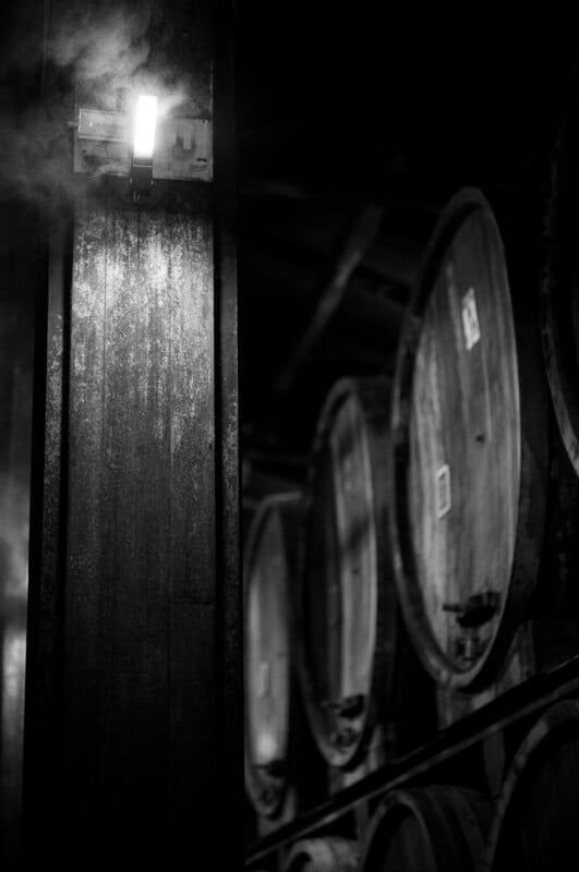 A dimly lit cellar with large wooden barrels stacked along the wall and a glowing light fixture on a pillar, emitting a small plume of steam or smoke in the dark atmosphere.