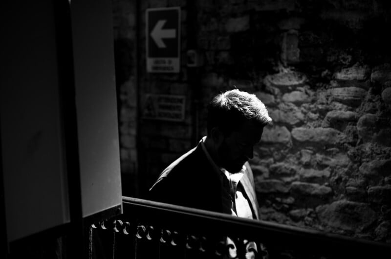 A man in a suit stands in a dimly lit area with a stone wall behind him. Only part of his face and upper body are illuminated, creating a dramatic shadow. A sign with a left arrow is visible in the background.