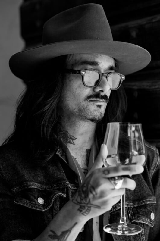 A person with long hair, a wide-brimmed hat, glasses, and visible tattoos holds a wine glass while looking down thoughtfully. The photo is in black and white.