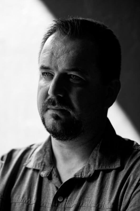 Black and white portrait of a man with short hair and a goatee, wearing a button-up shirt. He looks slightly to the side with a serious expression, illuminated by soft light from the left.