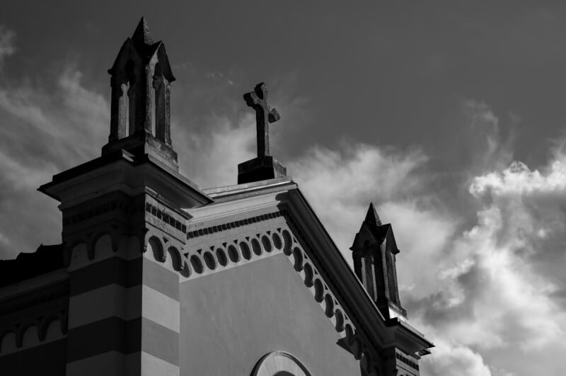 Black and white photo of a church roof with two small pointed towers and a central cross against a sky with scattered clouds. The building features decorative trim and arched details.