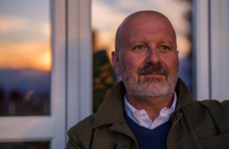 A middle-aged man with a beard and mustache sits in front of a window at sunset, wearing a collared shirt, sweater, and jacket, looking thoughtfully into the distance.