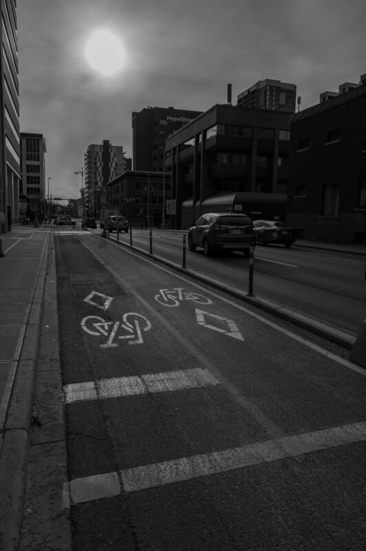 A black and white photo of a city street with cars driving, a separated bike lane marked with symbols, tall buildings on both sides, and the sun shining through a cloudy sky.