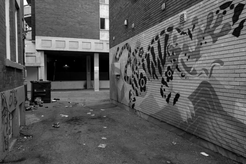 A black-and-white photo of a littered alley with graffiti art on a brick wall to the right, featuring abstract patterns. In the background, there's a dumpster and parts of several buildings.