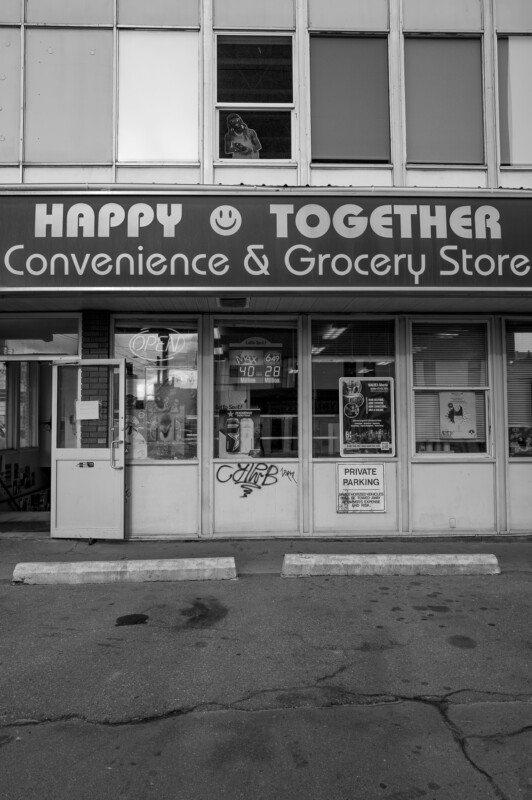 Black and white photo of "Happy Together Convenience & Grocery Store" storefront with signs, posters in the windows, and two empty parking spaces in front. A smiling face is part of the store's sign.