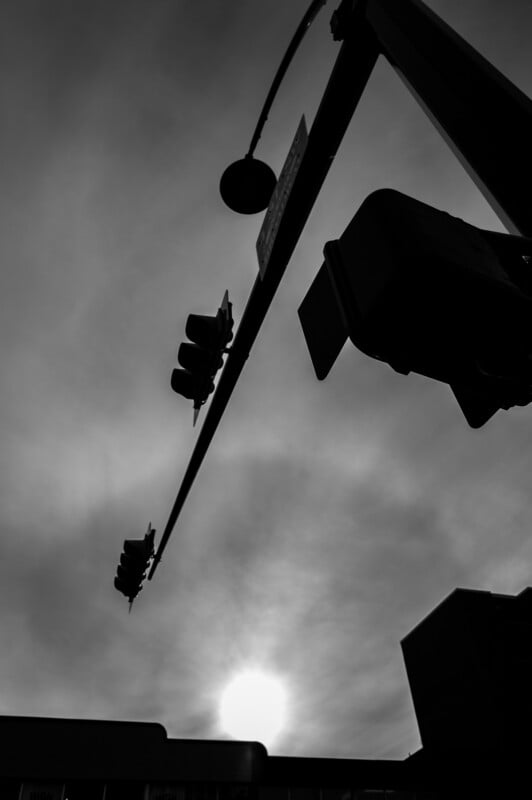A black and white photo of a traffic light and street signs silhouetted against a cloudy sky, with the sun low on the horizon and buildings faintly visible at the bottom.