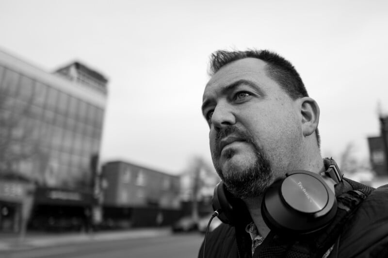 A man with short hair and a beard, wearing headphones around his neck, looks thoughtfully into the distance on a city street with buildings blurred in the background. The photo is in black and white.