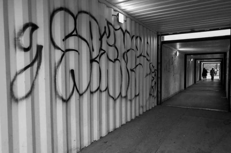 A black and white photo of a long, narrow corridor with graffiti on the left wall and two people walking away in the distance at the far end. The passage is lit by ceiling lights.