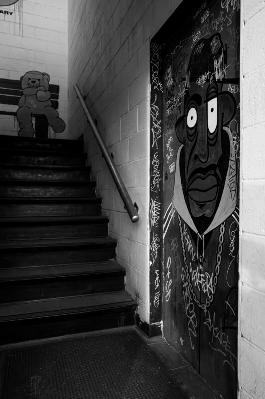 Black and white photo of a stairwell with graffiti; a cartoon face with a large chain is painted on a door, while a bear sits on a bench at the top of the stairs. Walls and door are covered with various tags.