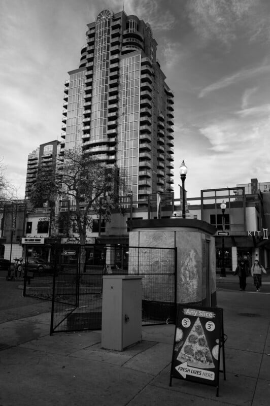 A tall modern apartment building towers over a city street. In the foreground, a sign advertises pizza slices; pedestrians and streetlights are visible. The scene is captured in black and white.