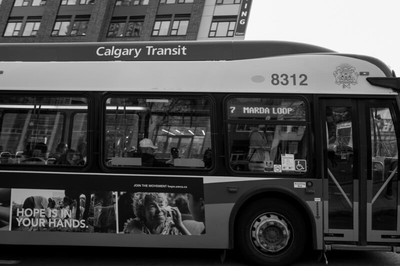 Black and white photo of a Calgary Transit bus with route 7 to Marda Loop. An advertisement on the side reads "HOPE IS IN YOUR HANDS" next to a smiling elderly woman. City buildings are visible in the background.