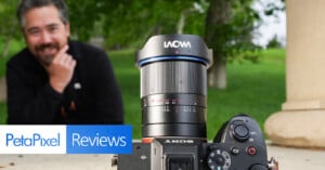 A camera with a Laowa lens is in focus in the foreground, with a smiling man sitting in the blurred background. The PetaPixel Reviews logo appears at the bottom left. The scene is outdoors with green trees and grass.