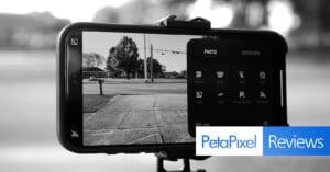 A smartphone mounted on a tripod is taking a black-and-white photo of a street scene. Camera settings are visible on the screen. The "PetaPixel Reviews" logo is overlaid on the bottom right corner.