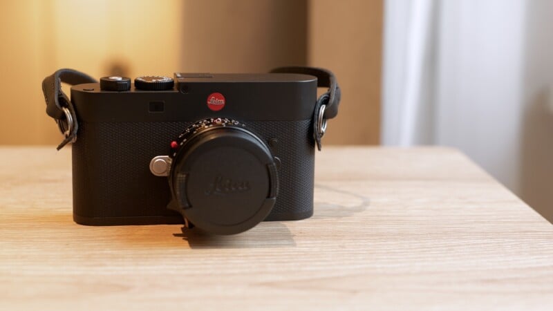 A black Leica digital camera with a lens cap sits on a light wooden surface, with a blurred background featuring warm lighting and a white curtain.