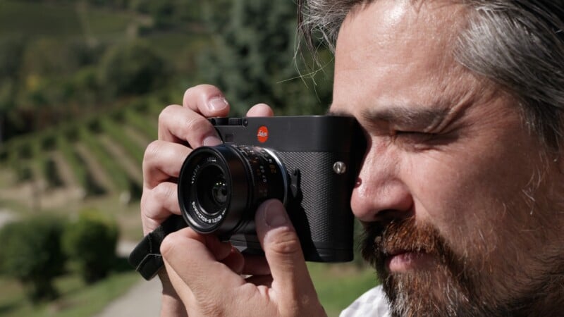 A man with gray hair and a beard is outdoors, holding a black Leica camera up to his face and looking through the viewfinder, with blurred greenery and vineyard rows in the background.