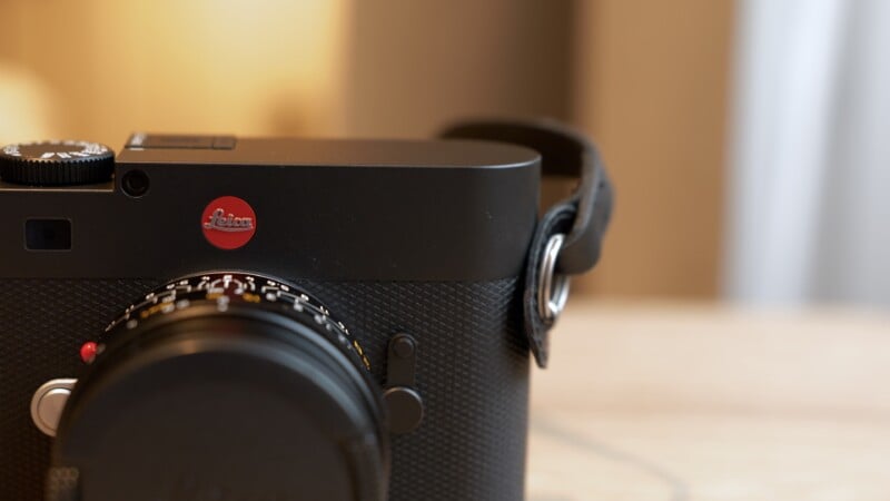 Close-up of a black Leica camera with a red Leica logo, focusing on the upper left side and lens, resting on a light-colored surface with a blurred background.