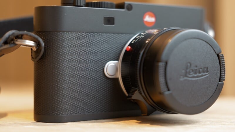 A close-up photo of a black Leica camera placed on a wooden surface, showing the textured grip, lens cap with the Leica logo, and part of the strap.