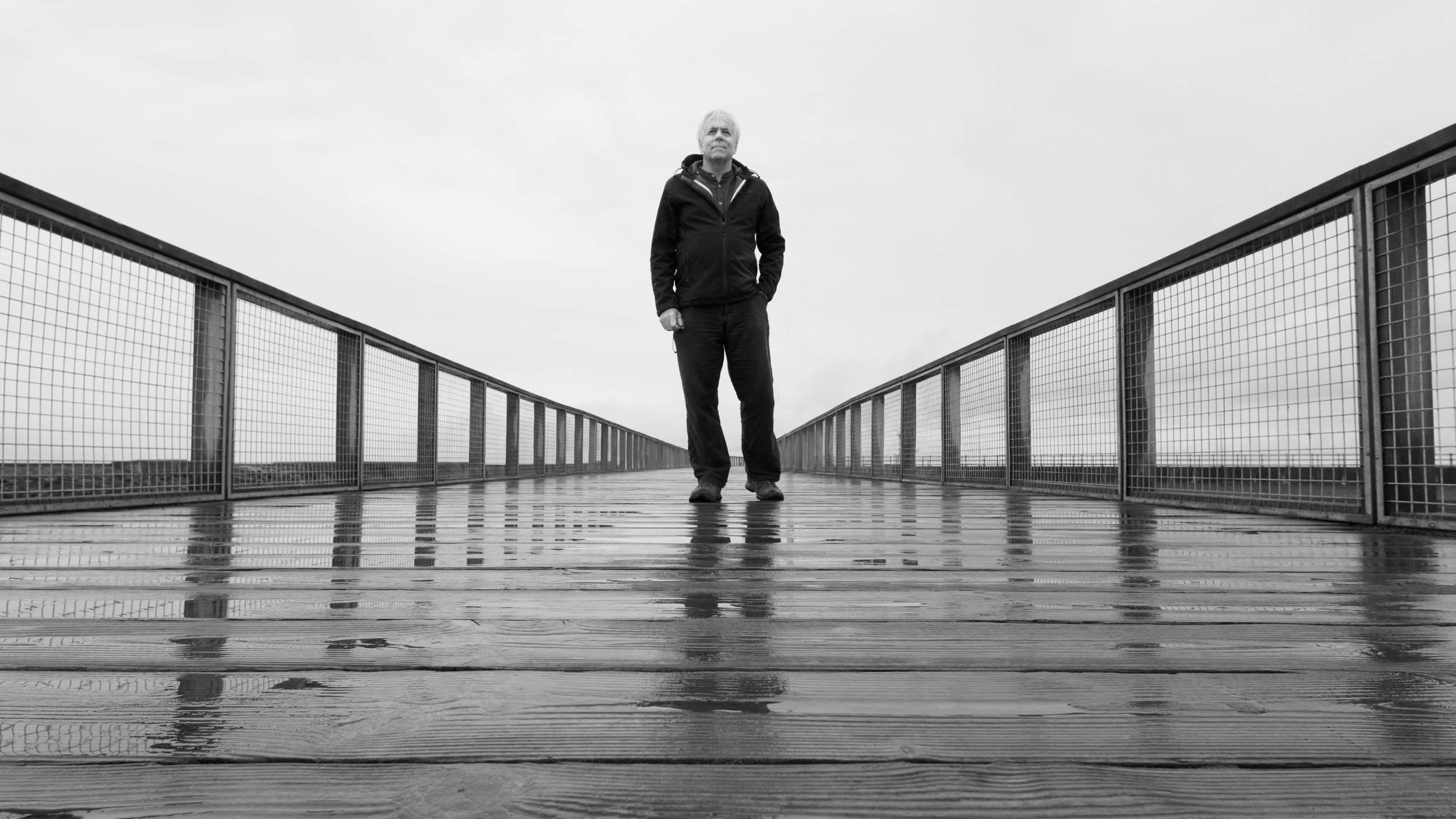 A man stands alone on a wet wooden pier with railings on both sides, looking ahead. The photo is black and white, with a cloudy sky in the background and reflections visible on the pier's surface.