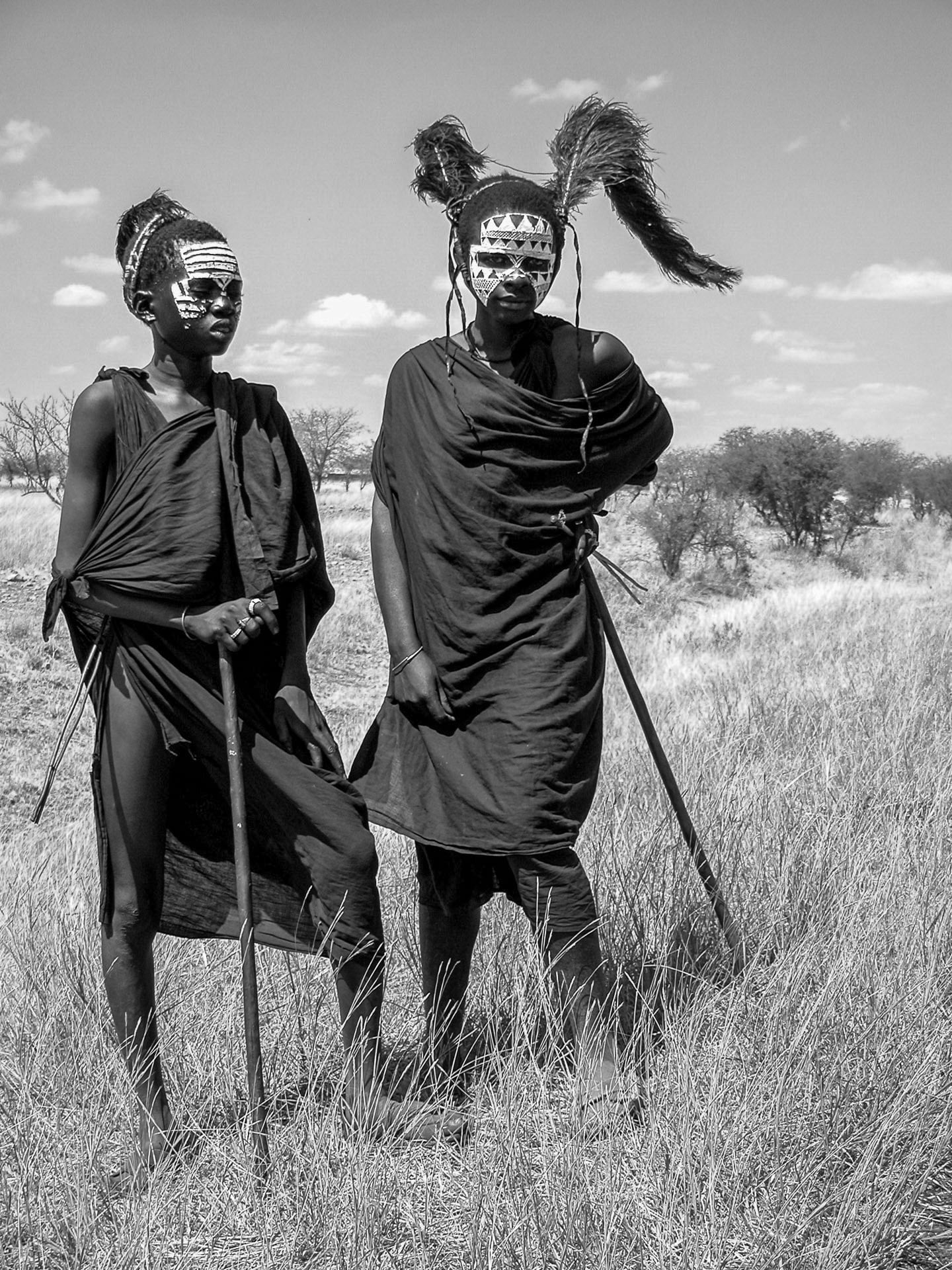 Two people stand in tall grass wearing dark traditional garments, with white painted patterns on their faces and elaborate feathered headpieces, each holding a stick, against a backdrop of trees and a partly cloudy sky.