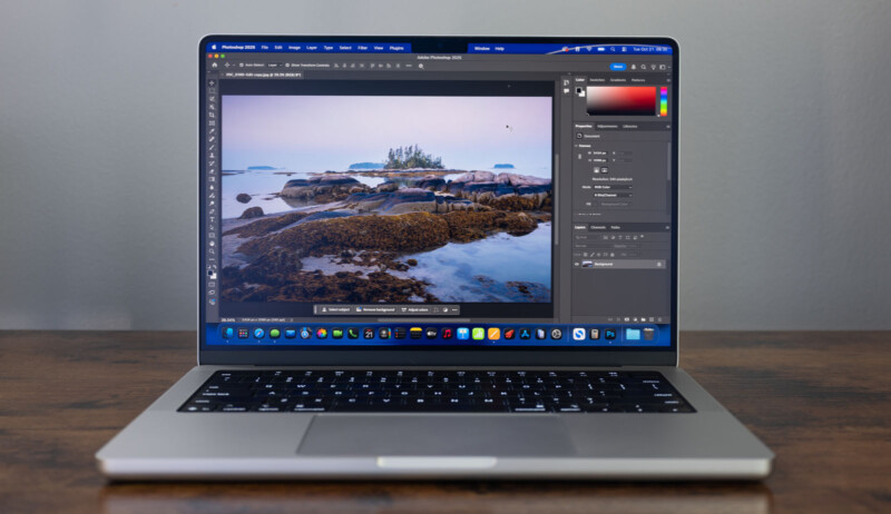 A laptop on a wooden table displays a photo editing software with an image of a rocky shoreline and small island, showing editing tools and adjustment panels on the screen.