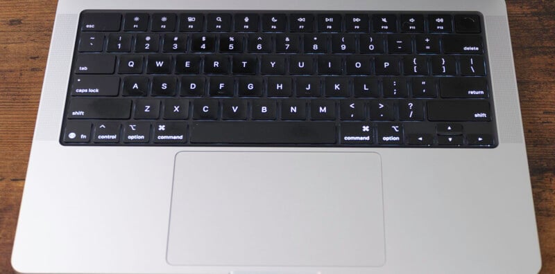 A backlit MacBook keyboard on a silver laptop, viewed from above on a wooden surface, with the trackpad visible below the keys.
