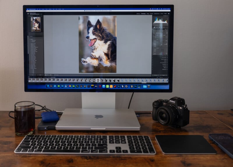 A desktop setup with a monitor displaying a photo editing program featuring a dog photo, a MacBook under the monitor, a keyboard, graphics tablet, digital camera, external drive, and a glass of dark beverage on a wooden desk.