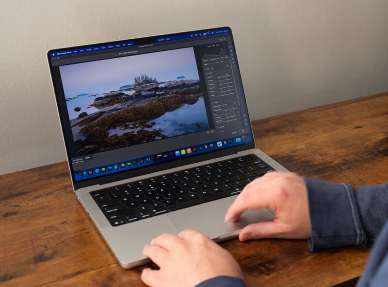 A person uses a laptop on a wooden desk, editing a landscape photo of a rocky shoreline with trees using photo editing software. The screen shows adjustment tools and a preview of the image.