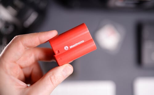 A hand holding a red Manfrotto camera battery with a blurred background of electronic devices and accessories.