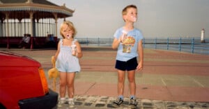 Two young children with ice cream on their faces stand on a seaside promenade. The girl holds a stuffed toy and wears a light blue romper; the boy wears a blue T-shirt and shorts. A red car and a lighthouse are in the background.