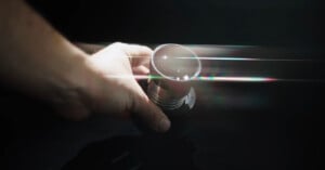 A hand holding a metallic cup against a dark background, with beams of light and subtle rainbow reflections passing through the scene.