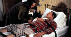 A woman leans over a man who is lying in bed, strapped down at the chest and legs with leather restraints, in a dimly lit, old-fashioned room with floral wallpaper.