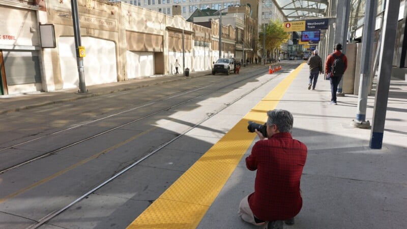 A person in a red plaid shirt kneels on a sidewalk, photographing a quiet city street with streetcar tracks. Several people walk in the distance, and a "Westbound Trains" sign is visible overhead.