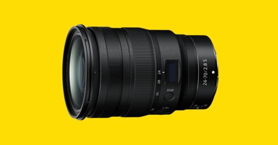 A black camera zoom lens with multiple control rings and a digital display, marked “24-70mm f/2.8 S,” is displayed on a bright yellow background.