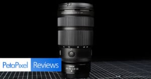 A Nikon NIKKOR 24-70mm f/2.8 S lens stands upright on a grid-patterned surface with a black background. The PetaPixel Reviews logo is displayed in the lower left corner.