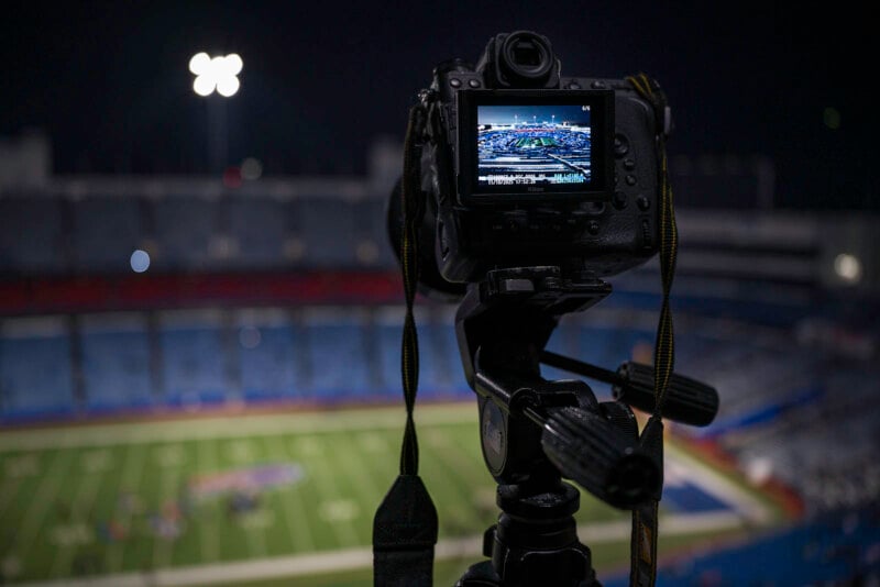 A camera on a tripod focuses on a brightly lit football field at night; the stadium appears empty in the background while the camera’s screen shows a clear view of the field.