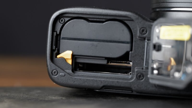 Close-up of an open camera battery compartment showing the battery and SD card slot, with a yellow latch securing the battery in place.