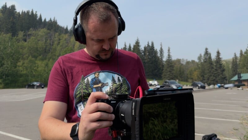 A man wearing headphones and a maroon shirt with a forest graphic operates a professional video camera outdoors, with trees and parked cars visible in the background.