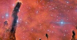 Tall, dark, pillar-like clouds of gas and dust stretch upward against a bright, reddish-orange nebula background dotted with numerous bright stars and glowing points of light.